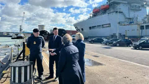 Three individuals in navy uniforms observe a large naval ship docked at a busy port. The individuals are dressed in navy uniforms which include dark jackets and trousers, with caps that add to the formal military look. In the background, the large aircraft carrier dominates the scene, its expansive flight deck and command tower standing out.