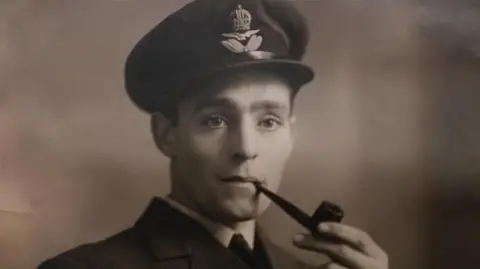 John Fairhall/BBC A black and white photograph featuring Roy Daines as a young man in military uniform. He has a smoking pipe in his mouth and he is looking past the camera.