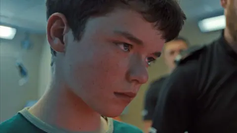 Netflix Owen Cooper, who plays Jamie, stands in a police station. He is staring off to the side. He wears a blue T-shirt and has short dark hair.