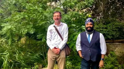 A little History of the Sikhs Stephen Barker and Rav Singh smiling for the camera outside. There is some greenery behind them. It's a sunny day.