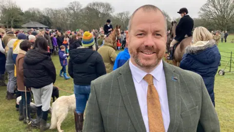 Will Day, joint Hunt Master, wearing a green tweed jacket and a yellow tie, stood in front of crowds of people and dogs and some people on horses.