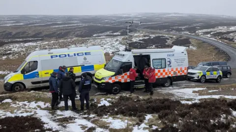 TWSMRT A police van parked alongside a mountain rescue van. Mountain rescuers can be seen in front of the vans in red jackets with police officers in black standing in a separate group. Moorland can be seen behind the vans, which stretches beyond the horizon.