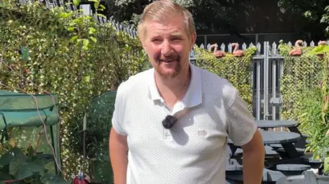 A man in a white T shirt stands in a garden