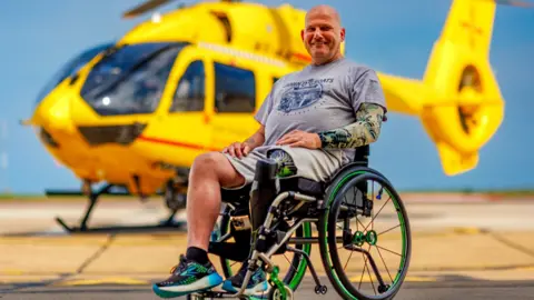 A man with an artificial left leg sits in a wheelchair wearing shorts and a T-shirt, with a yellow helicopter in the background.