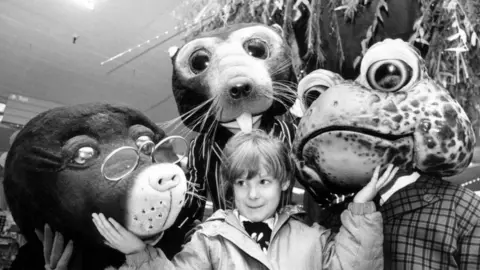 Getty Images A black and white photo of a young girl in a coast with her arms up under the chins of three large Wind in the Willows creatures