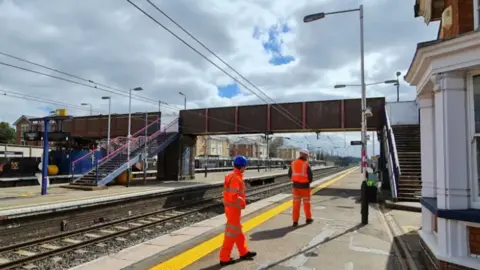 A close up of the old footbridge which is lacking paint and is no longer fit for purpose. Engineers in orange overalls and hard hats gather on one of the platforms.