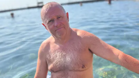 Sean Slattery - A man with a bald head, and his bare chest exposed in the water at the Bathing Pools. He has wispy blonde chest hair and is wearing blue and black swim shorts. 