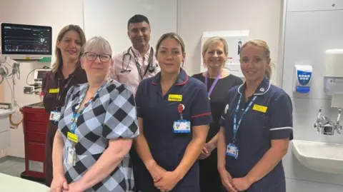 Six members of staff are standing in a line smiling at the camera. They are all wearing different medical uniforms and are standing in a cubicle on a ward. 
