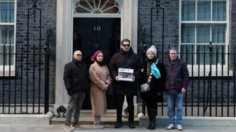 The group of five people stand wearing winter coats outside 10 Downing Street's door. The man in the middle who is wearing dark clothing is holding something that looks like a folder.
