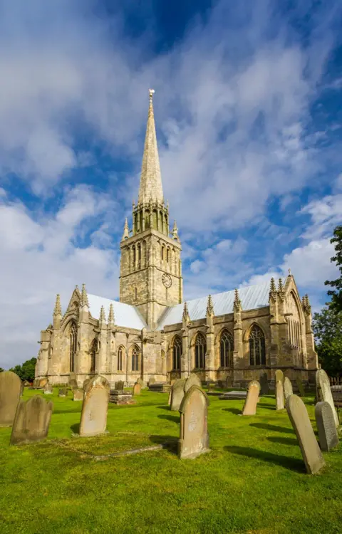 Universal Images Group/Getty Images An image of a large parish church built in the English gothic style with a tower and tall, graceful spire at its centre rising up into a blue sky. The stonework has a golden hue. In the foreground, gravestones stand in green grounds.