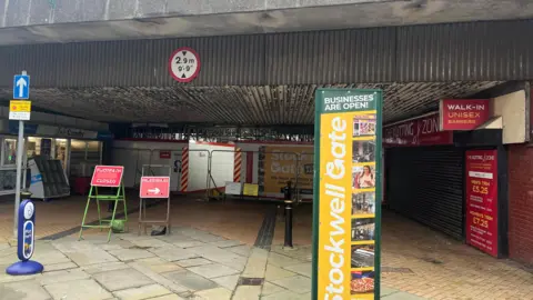 A pedestrianised street going underneath a bridge, with the far side sealed off with fences and hoardings