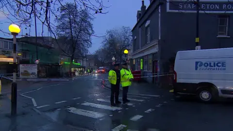 Two police officers stand on a zebra crossing with their backs facing a cordon at a murder investigation scene on a road lined with shops in Bristol. It is a grey and damp morning, which is still quite dark. A police federation van is parked beside them.