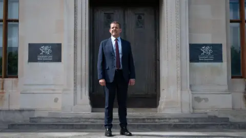 Welsh government The image, taken on Tuesday evening, shows Rhun ap Iorwerth standing on the steps of the Welsh Office. He is wearing a dark suit, white shirt, and red tie with white spots.
