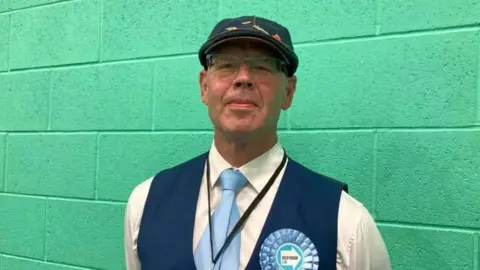 LDRS A man with a dark cap, glasses, a dark blue waistcoat, a white shirt, a blue tie and a light-blue Reform UK rosette, stands and smiles in front of a green-coloured wall.