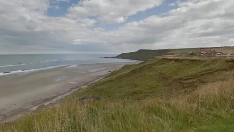 Shot of Cayton Bay, seen from the land - looking out to sea. The picture is taken on a grassy verge which sweeps down to the beach. Taken on a grey day.