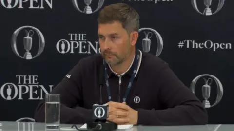 Chief executive of the R&A Mark Darbon is sitting at a microphone and has a glass of water nearby. He is in a black jacket with a blue lanyard on. He is standing in front of black wall with The Open insignia visible