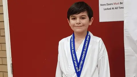 Cotter Family Ben Cotter when he was a young boy, wearing a karate gi and with a medal around his neck. He is smiling into the camera.