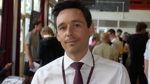 Jamie Niblock/BBC A man wearing a white shirt and a tie. He is also wearing a black lanyard and has short dark brown hair.
He is standing in a school and has a lot of people behind him 