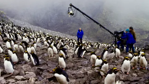 Ben Osborne The image shows a film crew working among a vast colony of penguins on a rocky, mist-covered hillside. A large camera crane extends over the scene while crew members stand and operate equipment, surrounded by hundreds of penguins clustered across the terrain.