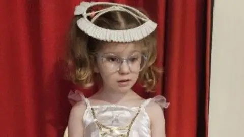 Cancer Research UK Ella is wearing plastic-framed spectacles. She is wearing her blonde-brown hair in two pigtails with a halo on her head, and a shimmery ivory strappy dress with gold asymmetric lines on the front.