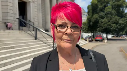 A woman looks at the camera. It is a head and shoulders shot and she has a black blazer on. She has glasses on and short bright pink hair.