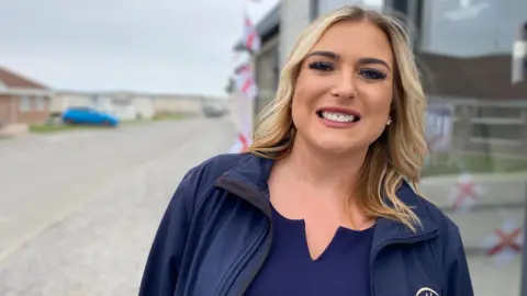 Louise is smiling in front of a caravan at the park, wearing a branded blue jacket and top. She has blonde shoulder length hair