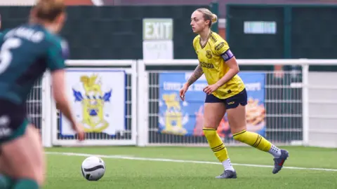 Lucy Copsey A female footballer wearing a yellow and navy blue football kit and Nike football boots mid run with the ball. She is wearing a captain's armband and looking up in the distance. 