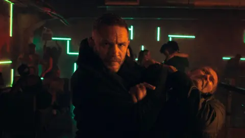 Netflix Tom hardy, who has another man in a chokehold. Both men are wearing all black and in the background is a nightclub with people dancing.