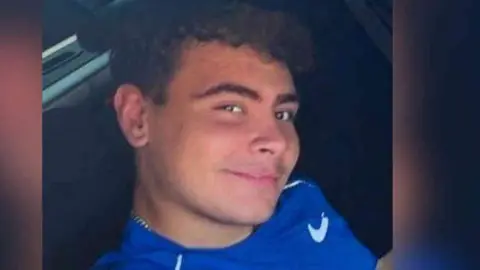 A teenager with curly brown hair and brown eyes smiles at the camera. He is wearing a blue top with a white trim and a branded tick. 