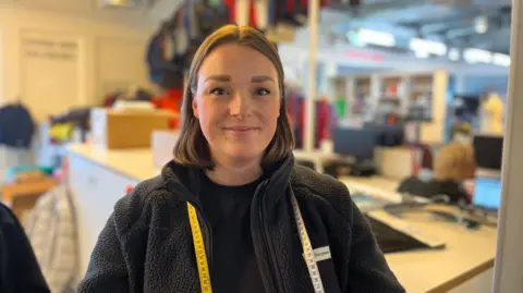 Femeke Vesters stands in front of a Berghaus production line filled with coats. She is wearing a fleece with a tape measure round her neck. 