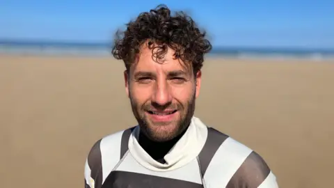 A man stands on Watergate Bay beach, it is a bright sunny day and the sky is very blue. He is wearing a wet suit with a black and white striped rash vest over the top.