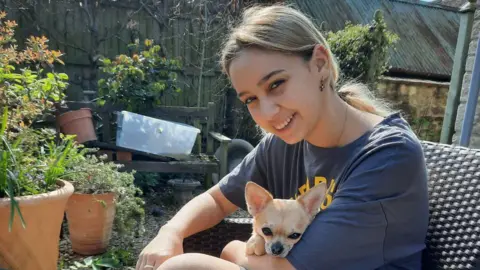 Viktoriia Izviekova Image shows Viktoriia sat in a garden with her Chihuahua. Behind her is a garden fence. There are lots of plants in brown pots stood in front of it as well as a wooden bench. Viktoriia is sat on garden chair with her Chihuahua on her lap. Her chihuahua is beige in colour. Viktoriia has her hair tied back in a low pony tail. She is wearing a blue oversized shirt with yellow writing on. She is smiling straight at the camera. 