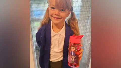 Family photo Ruby has long blonde hair in bunches. She is smiling widely at the camera and is wearing stud earrings, white polo shirt, blue school cardigan and grey skirt. She is holding a red water bottle, printed with a cartoon version of her image and her name on it.