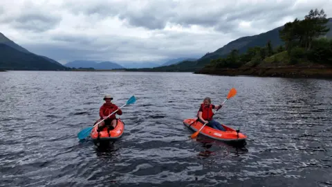 Rob Kingsland and Lorna Gordon paddle in the loch as they discuss tourism - the kayaks are bright orange and they both wear red life vests. The scene behind them is very picturesque.