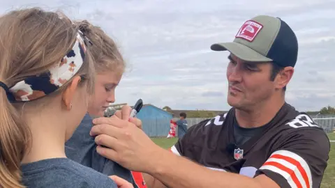James Buriz/BBC Gary Berniz é um estudante de duas em idade principal. As meninas estão olhando para o olhar, o cabelo justo está puxando atrás da cauda do pônei e usando uma camiseta cinza. Gary Berniz está à direita, barbeada limpa, um boné de beisebol azul e verde claro e uma camiseta marrom. Ele está assinando a camiseta de uma garota. Eles estão no playground. 