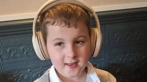 Rhys with his mouth open in a slight smile. He has short, light brown hair and is wearing large cream-coloured headphones over his ears. He is looking to his right and is wearing a grey and black polo shirt.