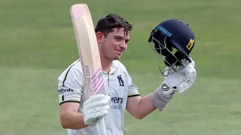 Ethan Bamber raises his bat and helmet to celebrate his century for Warwickshire