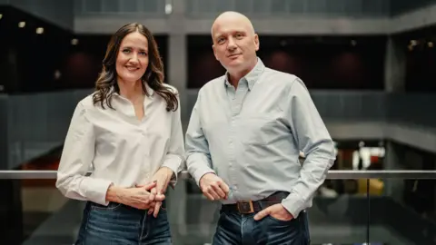 BBC Scotland Laura Maciver and Martin Geissler casually lean on a glass railing inside the BBC Scotland building. They are both wearing jeans. Laura has long, dark hair and is wearing a white shirt. Martin has a bald head and is wearing a light blue shirt.