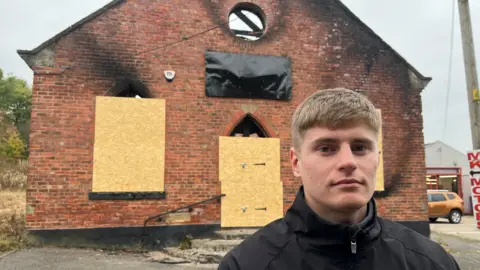 Kai Richmond is standing in front of a the burnt-out brick gym, which has boarded windows.