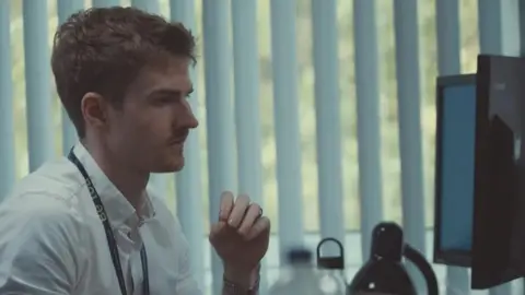 Channel 4/24 Hours in Police Custody A man wearing a white shirt with short hair is sat at a desk and looking at a computer screen