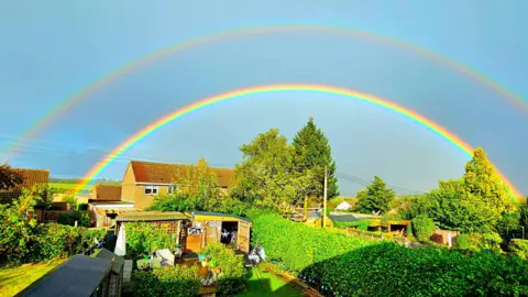 PennyB The sun is shining after heavy rain across a garden seen with neighbouring houses in the background. Above it all shine two full rainbows.