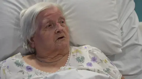 The image shows an elderly woman lying propped up in a hospital bed, resting against large white pillows. She is wearing a light‑coloured floral hospital nightdress and is covered by white bedding pulled up to her chest. 