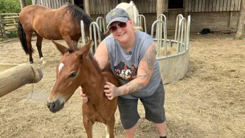 A woman, wearing sunglasses and a cap, smiles at the camera. She has her arms around a brown foal. There are two horses feeding from a trough in the background, with a barn behind them.