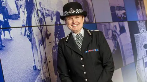 Durham Police Rachel Bacon is smiling as she stands next to a wall covered in purple-tinted photos. She is wearing a police uniform, including a hat. 