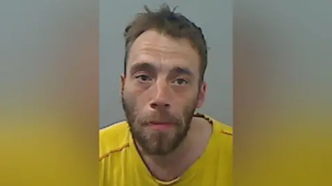 Cleveland Police mugshot of a man with a brown beard and curly hair, wearing a yellow T-shirt.