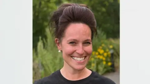 Natasha Streatfield Natasha Streatfield is looking towards the camera smiling. She has brown hair which is up in a ponytail, and is wearing a black T-shirt. In the background are flowers and trees which are blurred.