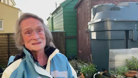 BBC Pensioner Pauline Trubody stands in her garden next to a dark green heating oil tank, which is next to a brown shed. She has long grey hair and is wearing a blue and white coat.