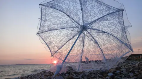 Stacey Johns An umbrella on a pebbly beach with the sunsetting in the distance on the water visible through the transparent umbrella. There are people in the distance by the water.