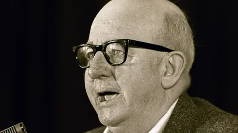 Pacemaker A black and white image of Joe Cahill. He is bald and wearing glasses speaking in front of a microphone.