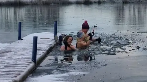 Kellie Tarr A group of women in bikinis using hammers to break a layer of thick ice covering a lake which they are in